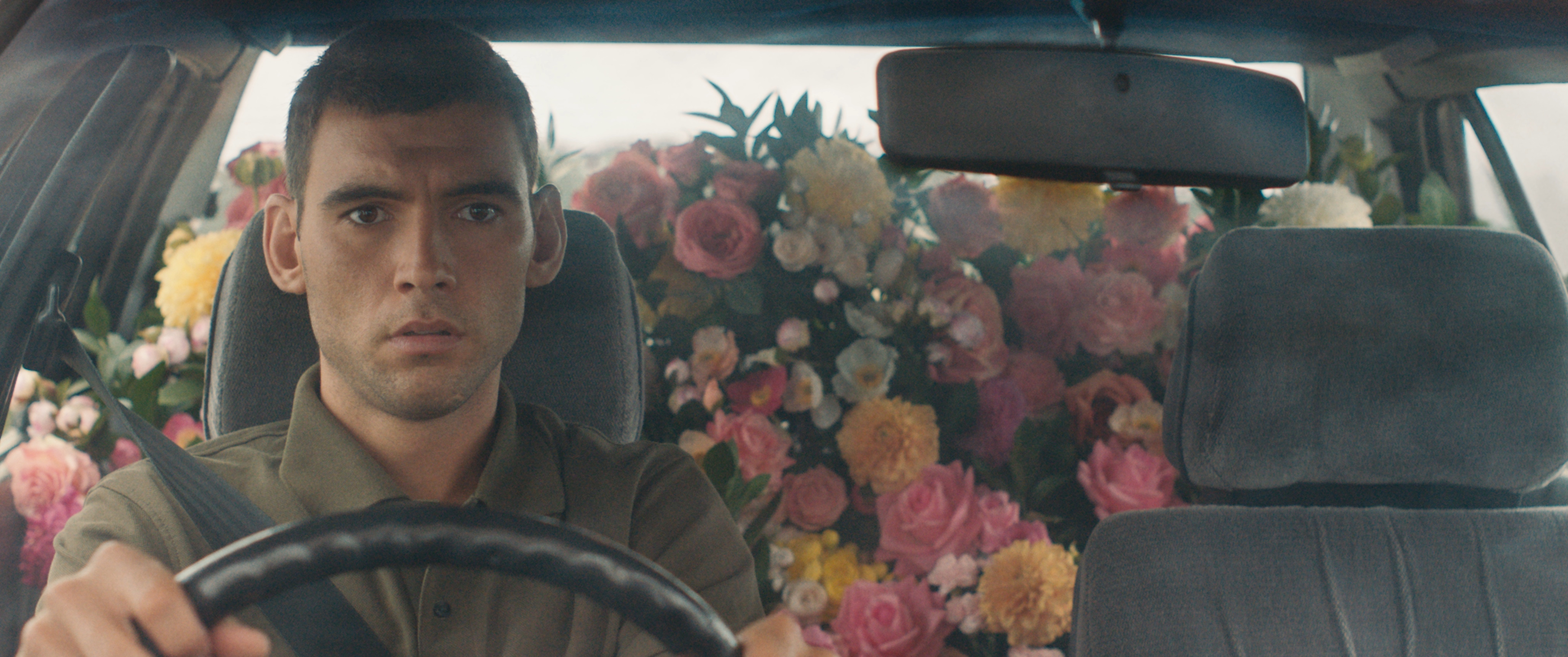 Film still of a flustered young man driving a car with a huge assortment of arranged flowers in pink, yellow, and white in the backseats.