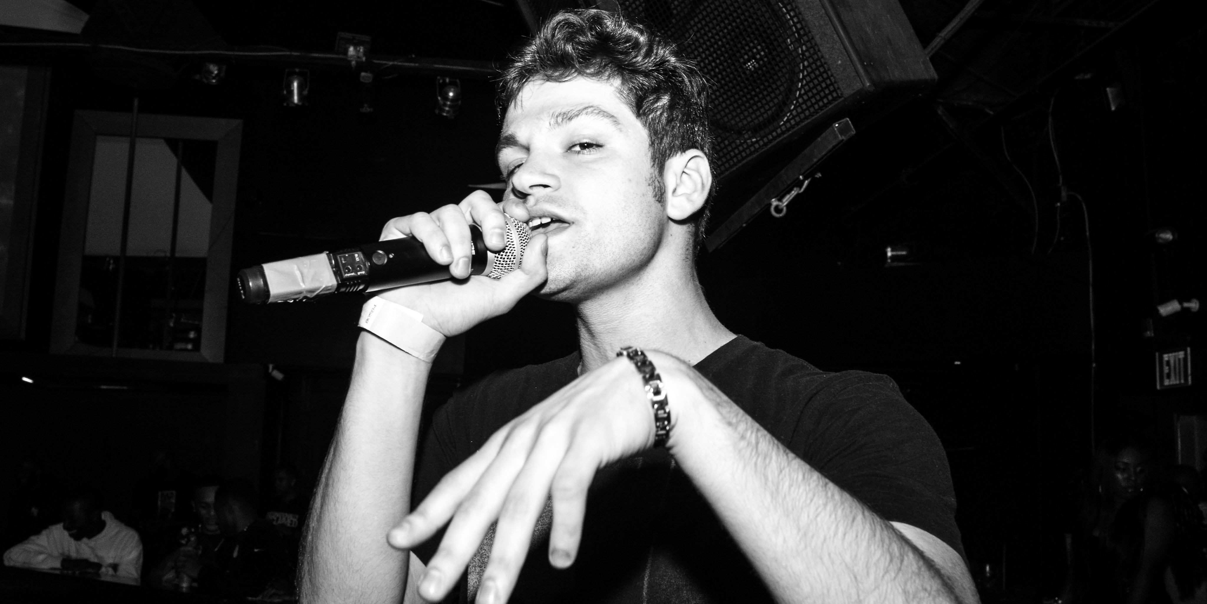 Black and white film still of a young man singing into a handheld mic, glancing at the camera with a confident expression.