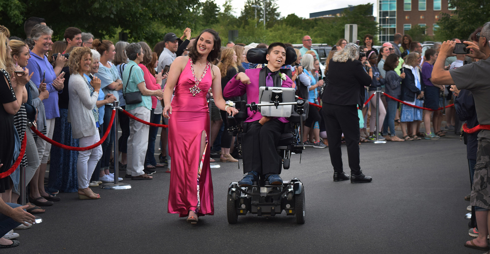 Wide film still of a smiling young couple dressed for prom, an ambulatory girl wearing a long pink dress, and a boy in a wheelchair wearing a purple tux. A large, cheerful crowd of people looks on from behind a red velvet rope barrier.
