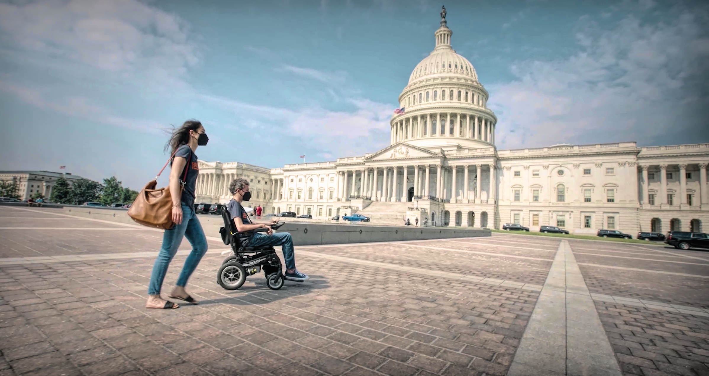 Wide film still of an ambulatory woman and a man in a wheelchair heading towards the U.S. Capitol, a cloudy blue sky above them.
