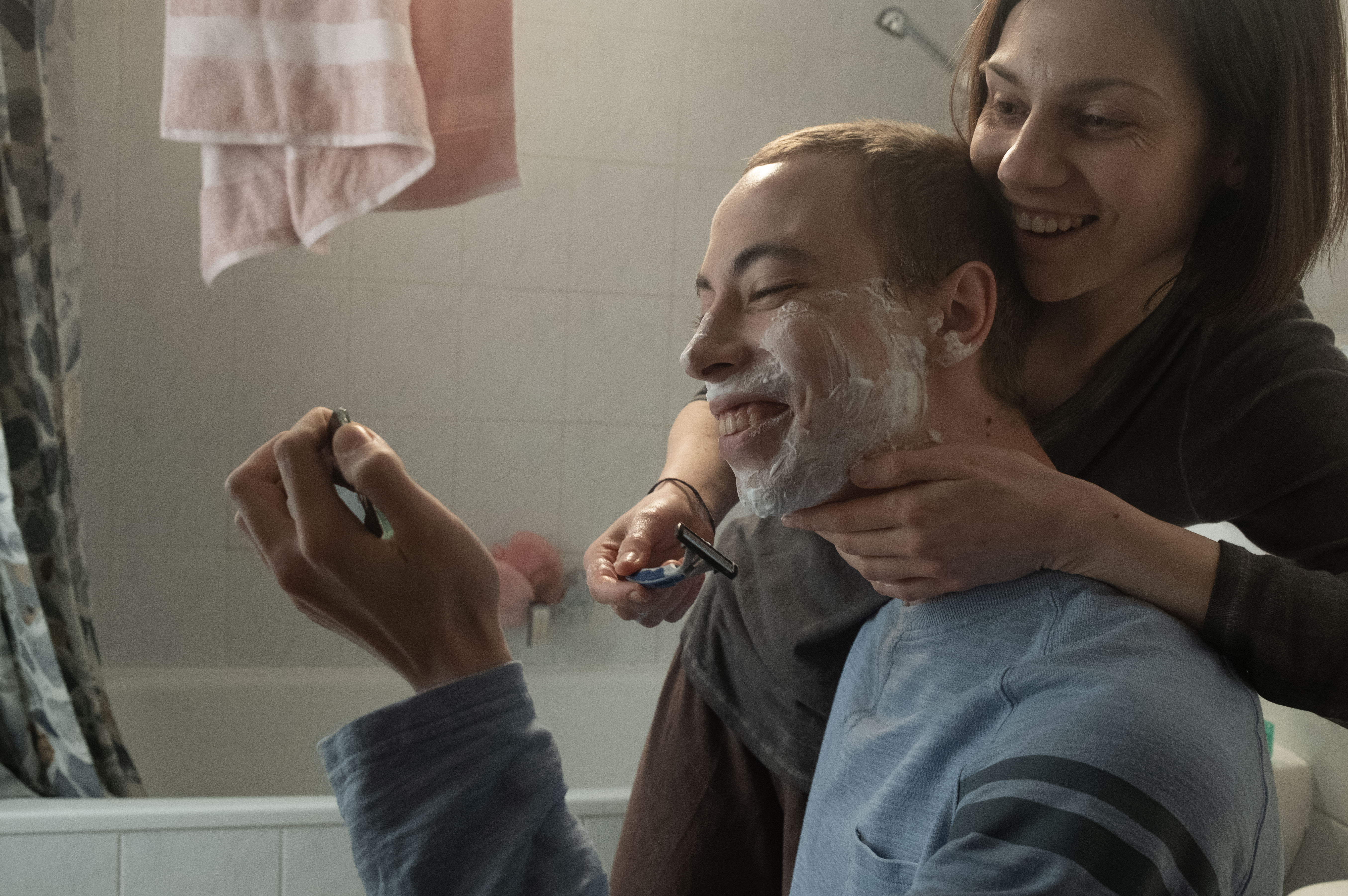 Film still of a smiling mother and her teenage son. The mother shaves her son’s face in the bathroom.
