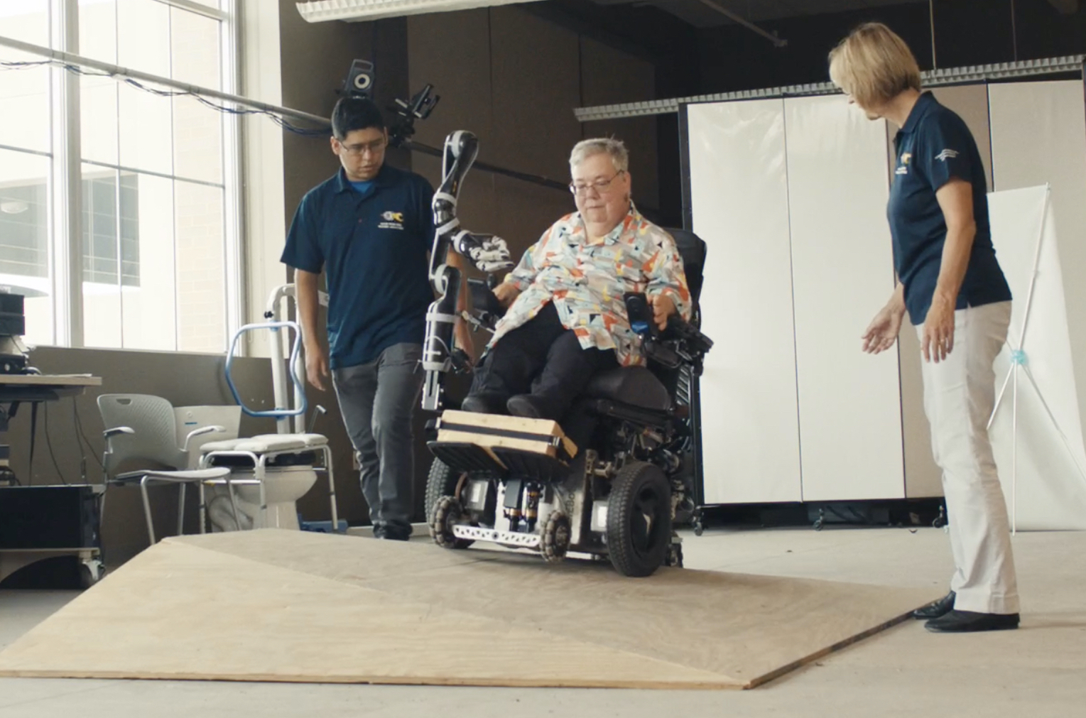Film still of an older woman in a high-tech wheelchair in a laboratory. She is driving over a wooden ramp with bumps and edges, designed to test the wheelchair. Two people in uniform polo shirts are watching her on either side.