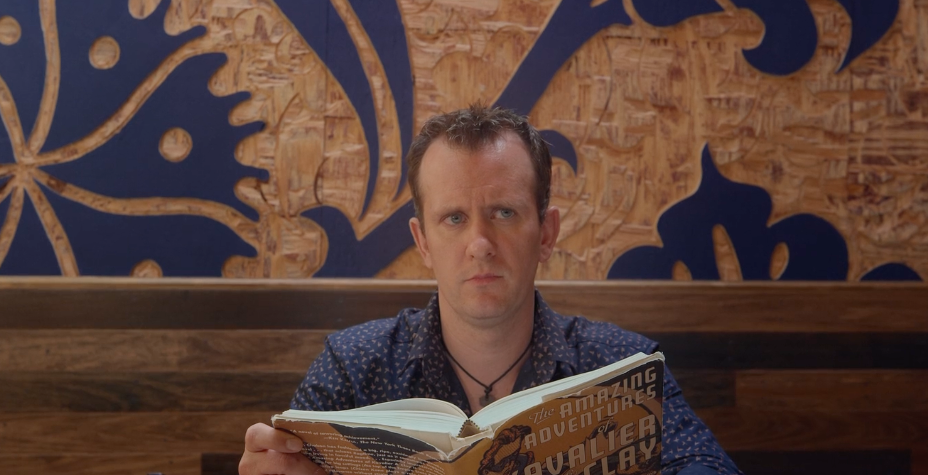 Film still of a man looking up from his book with a curious expression. He appears to be sitting in a booth in an earthy, eclectic café.