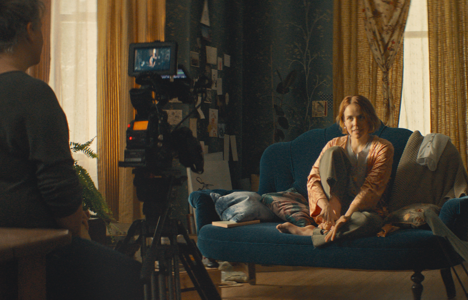Film still of a young woman sitting cross-legged on a velvet couch in a bohemian room, looking into a camera on a tripod pointing at her in the foreground. The camera operator’s back is partially in frame.