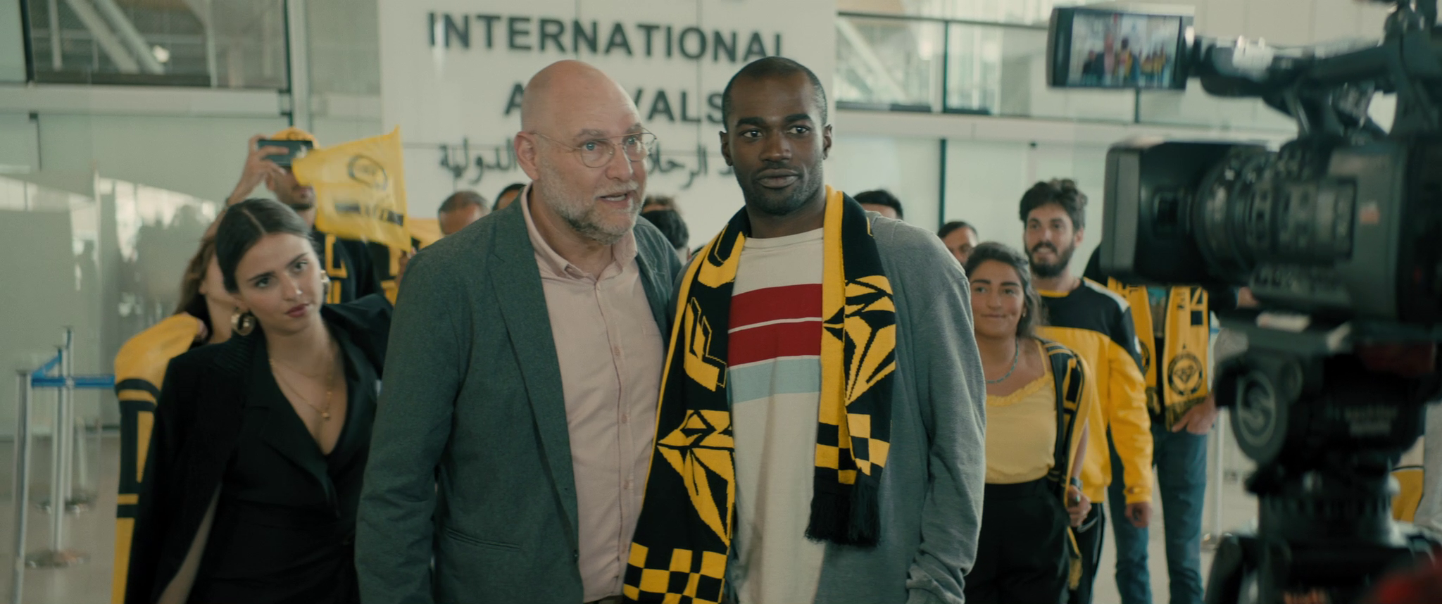 In an airport, a young Eritrean man with a black-and-yellow sports team scarf on his shoulders is surrounded by people and looks into a large professional video camera at the edge of the frame.