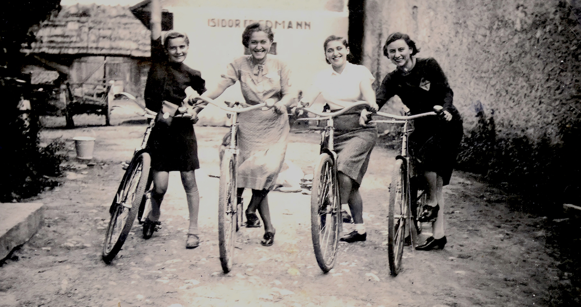 An old black and white photo shows four women on bikes, smiling happily and posing for the camera. They appear to be on a street in a town.