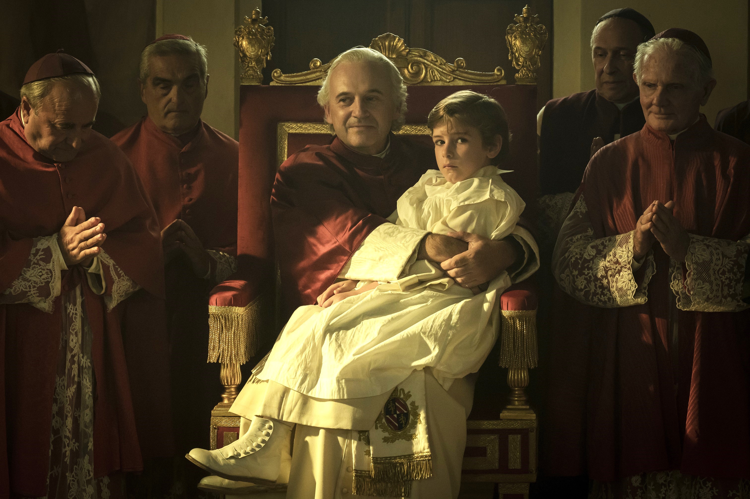 A young boy dressed in a white religious gown sits in the lap of the Pope, sitting on a throne. The two are surrounded by other clergymen.