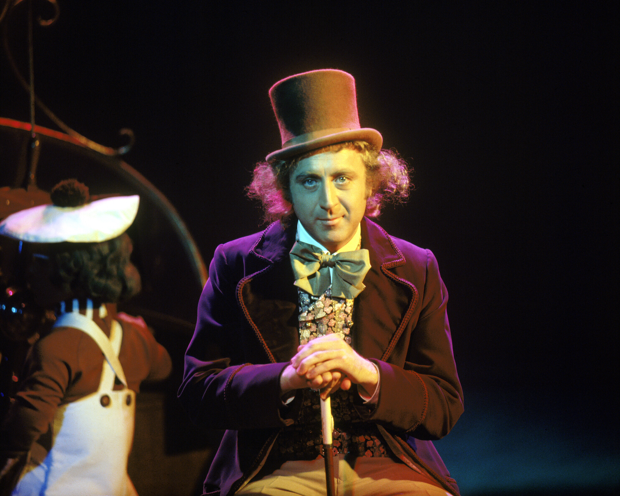 Gene Wilder, dressed in his Willy Wonka costume from Willy Wonka & the Chocolate Factory (1971), looks directly at the camera. An Oompa Loompa can be seen in the background.