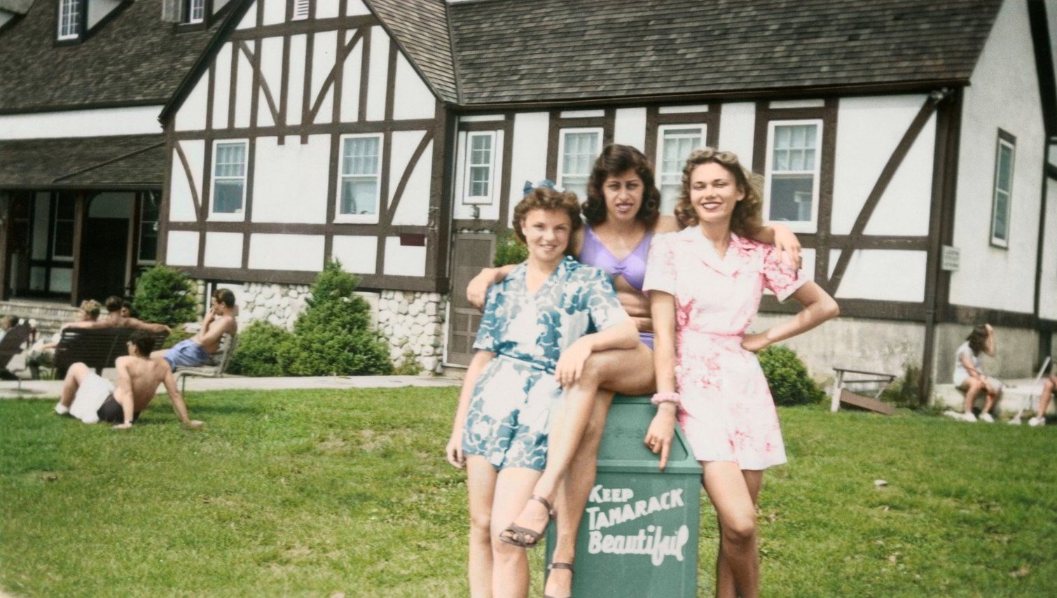 An old color photograph shows three women in brightly-colored summertime outfits smiling for the camera with their arms draped around each other, perched on a trashcan that reads “Keep Tamarack Beautiful.” Behind them is a summer resort with other vacationers dotting the area behind them.