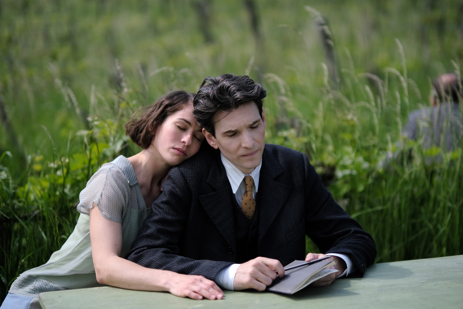 A woman lays her head on the shoulder of a man as they sit at a table near a field of tall grass. The man holds a pencil and small notebook.