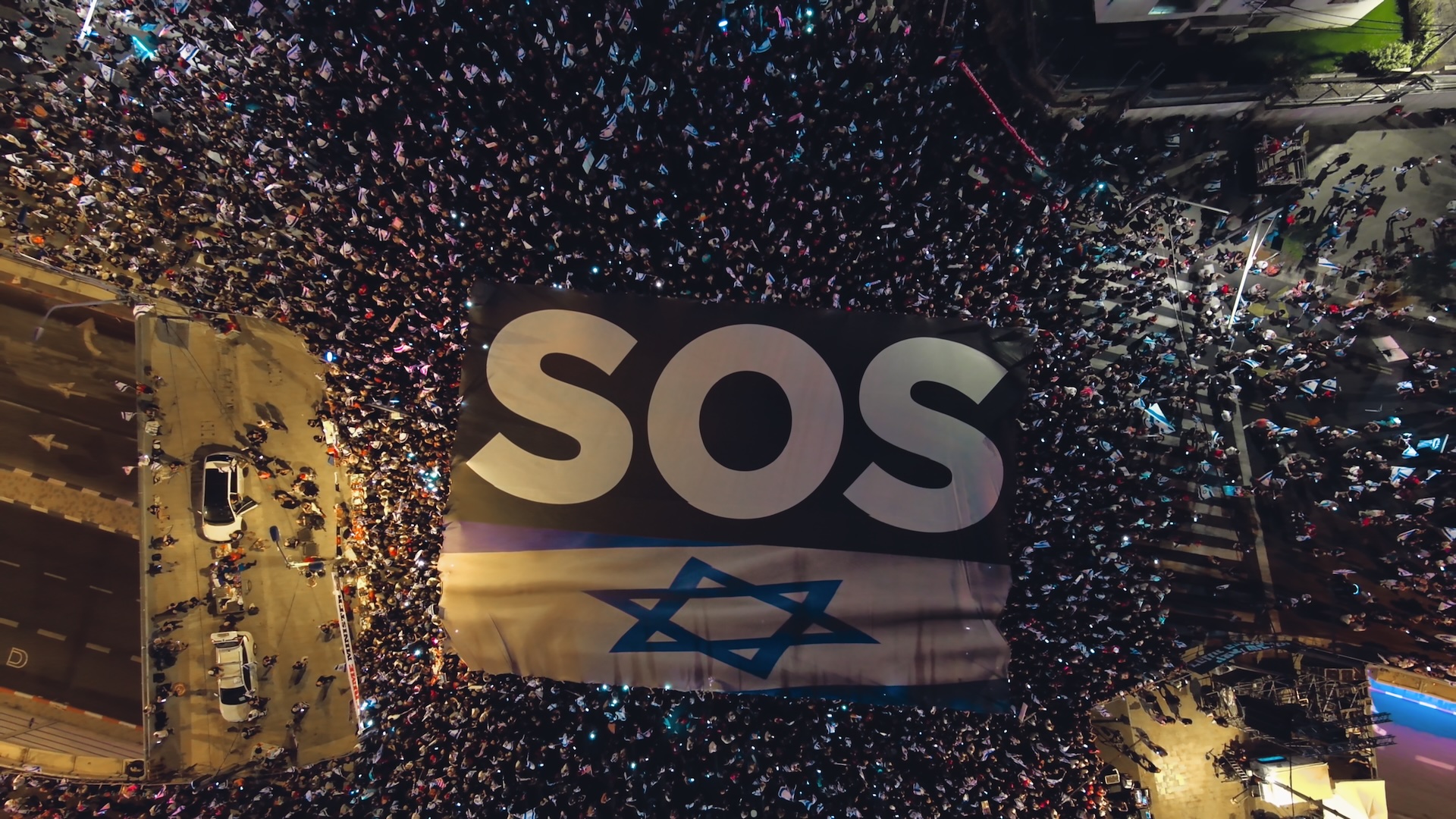 From above, a large crowd of people hold a giant banner with the phrase "SOS" and the Israeli flag.