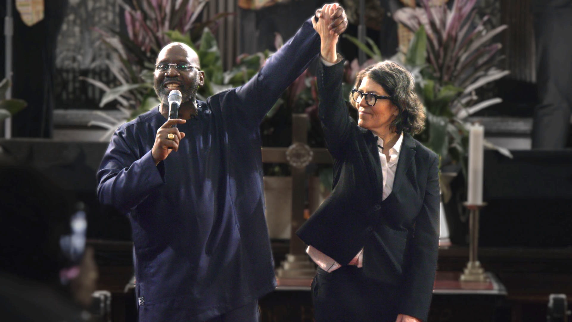 A black pastor speaks into a microphone while holding hands with a white Rabbi, who looks at him and holds their joined hands aloft.