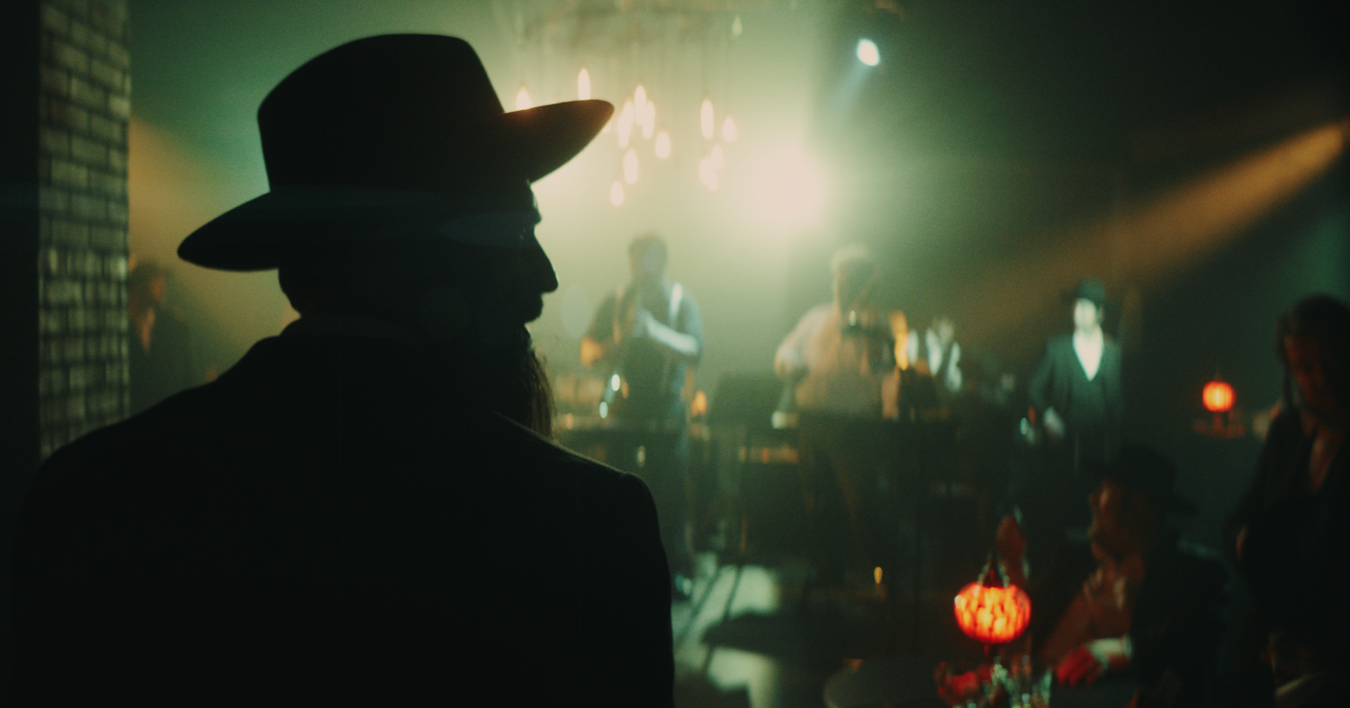 A silhouette of a bearded man wearing a wide brim hat in a dark room, where a few central lights illuminate several musicians and patrons.