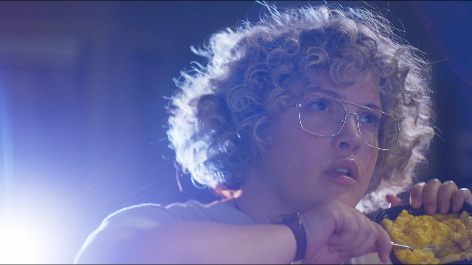 A young person with glasses stares intently with a projector flare behind them. They are holding a bowl of mac and cheese.