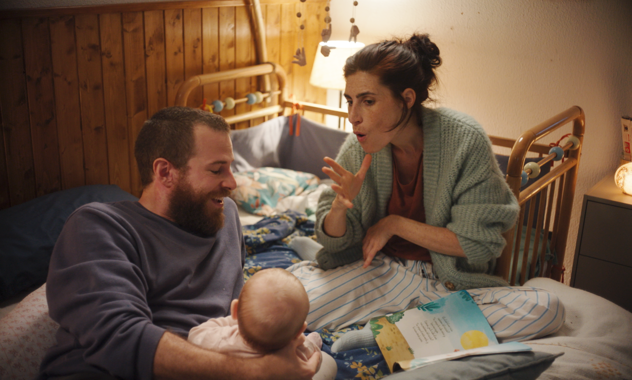 Film still of a mother signing to her baby while the father smiles, all sitting together on a bed in a softly lit nursery.