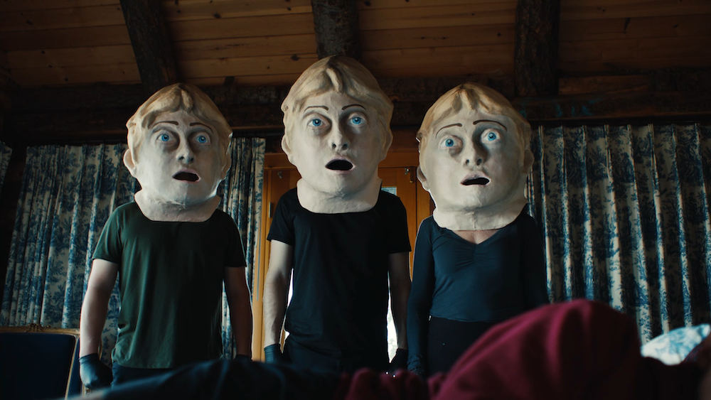A medium shot from the film “RAT” of three people wearing similar paper mache heads with blonde hair, blue eyes and an agog expression.