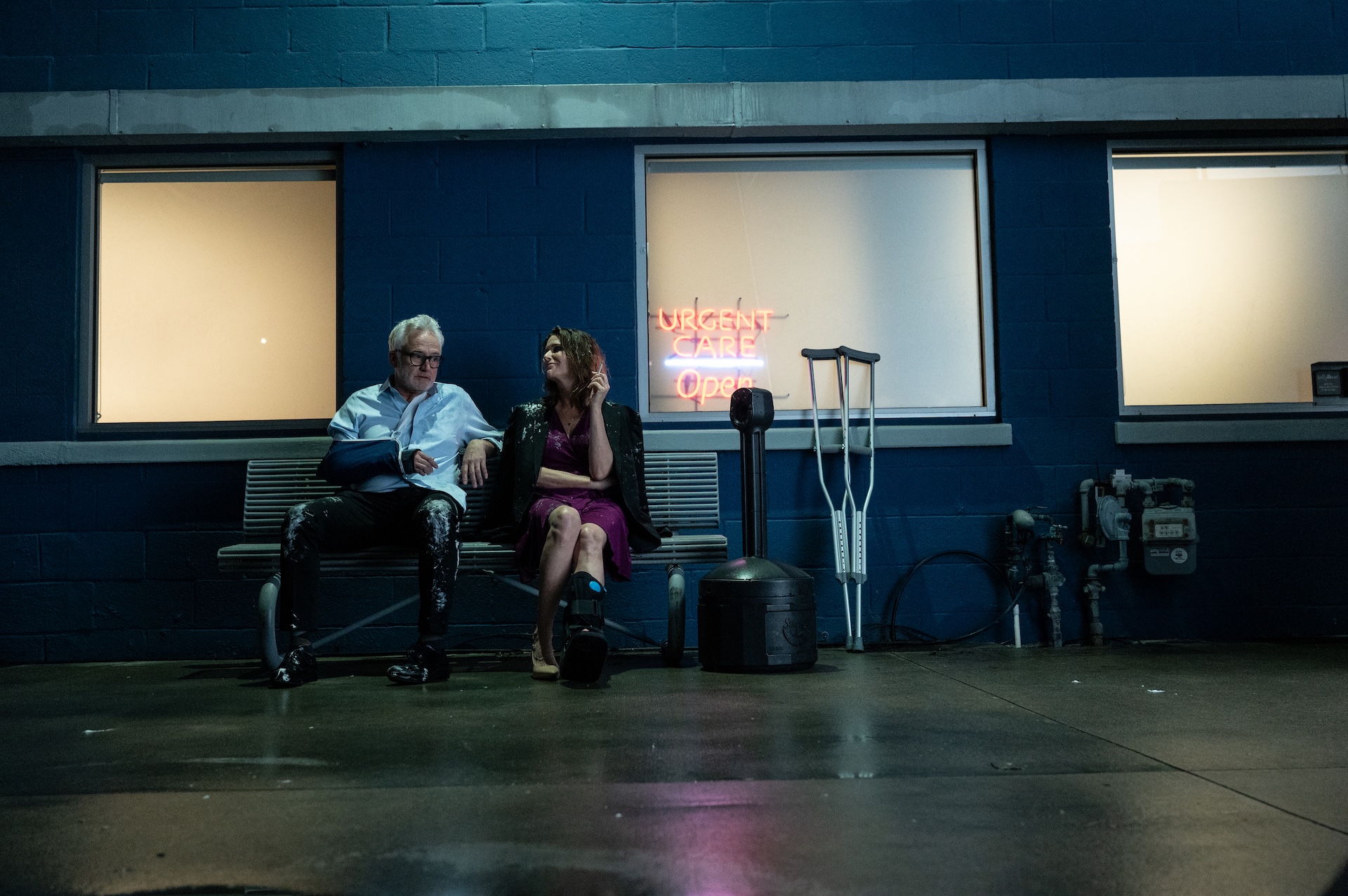 A wide shot from the film “For Worse” of an old man and a woman sitting on a bench. The man is wearing a cast and the woman is holding a cigarette. Next to them a pair of crutches stand.