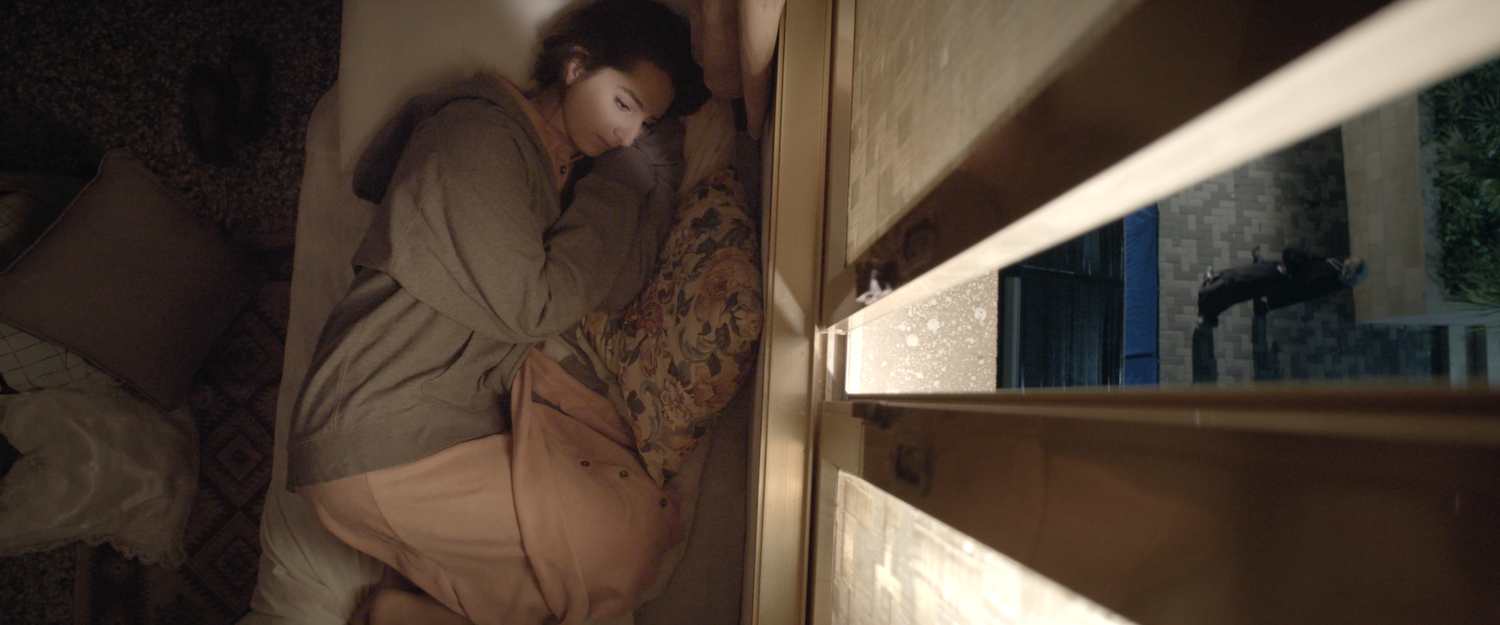 A medium shot from the film “The Source of All Beauty” of a woman laying in bed next to a slightly open window.