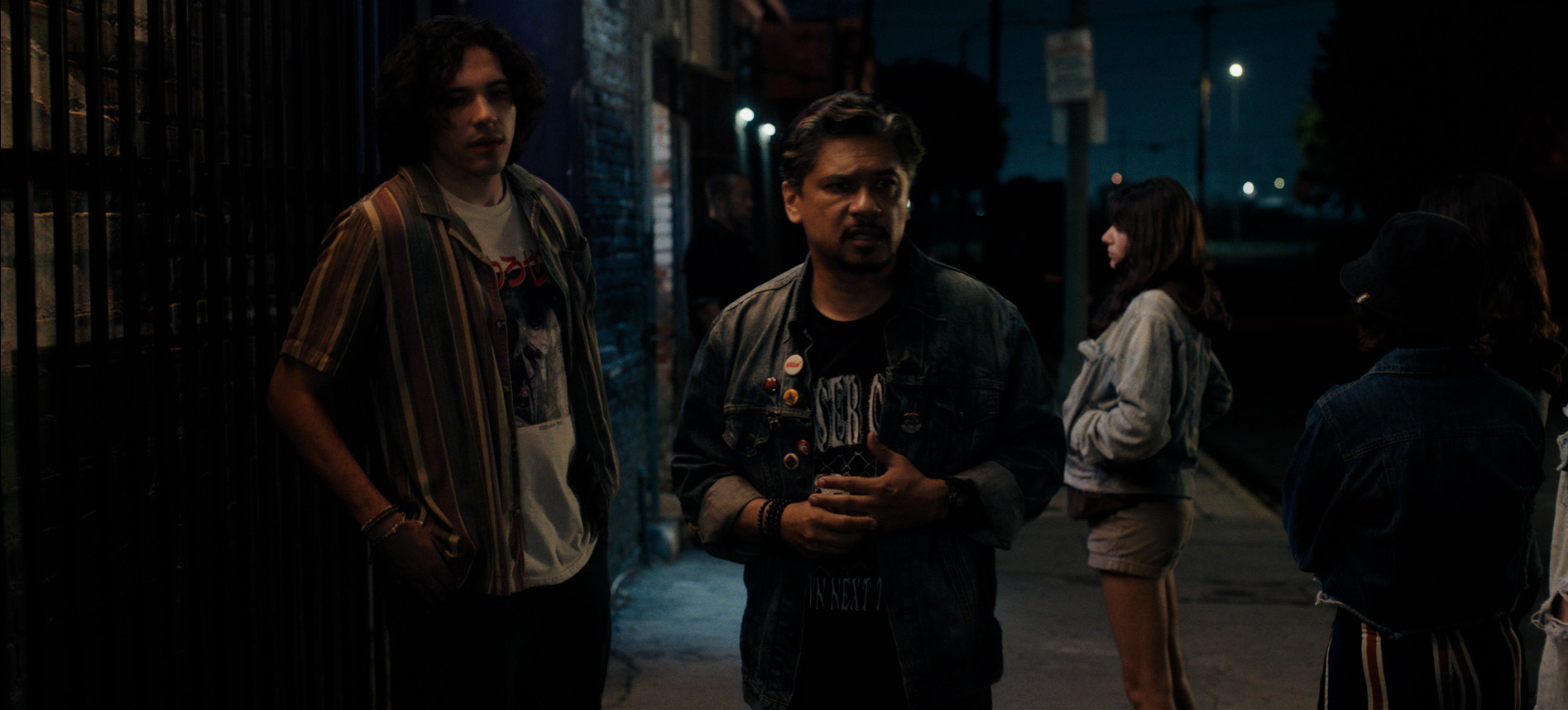 A wide shot from the film “Mosh” of two men standing on the sidewalk. The building in the background is the only light source in the dimly lit street.