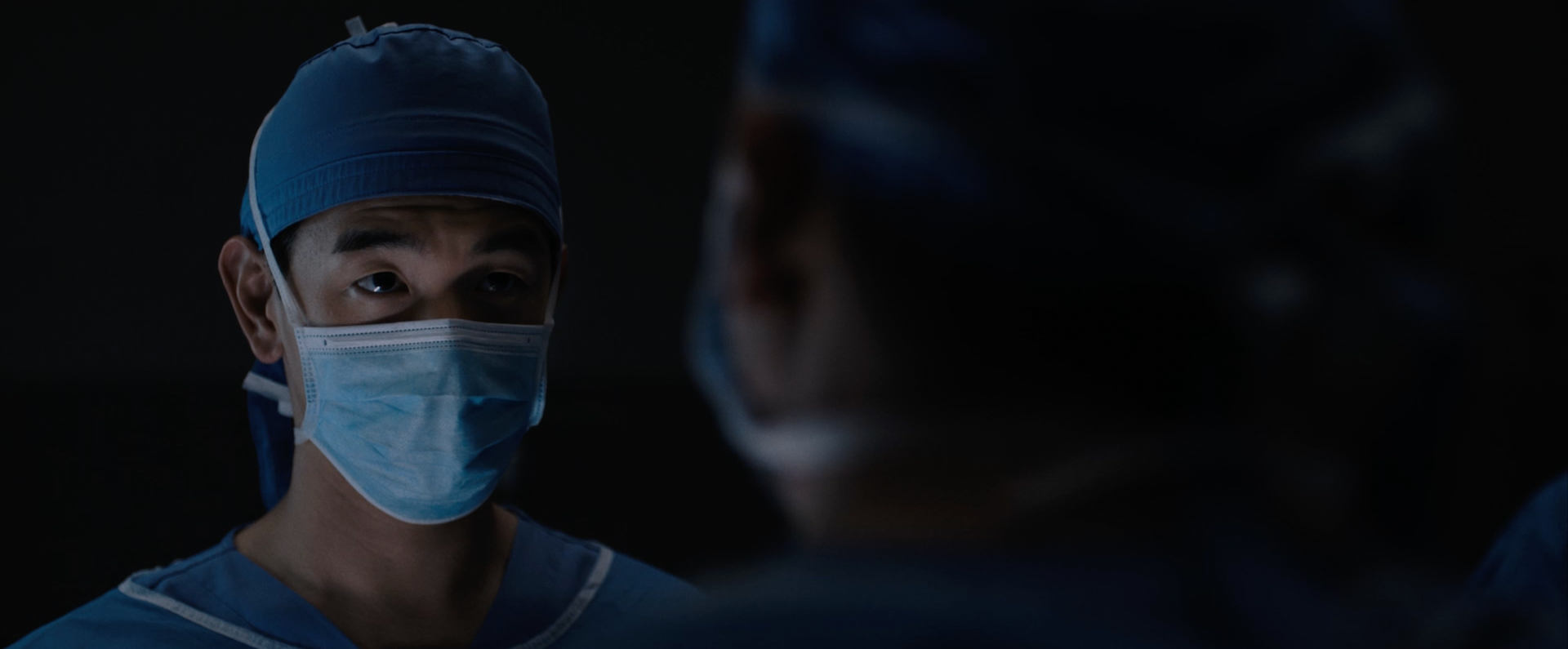 A medium shot from the film “Transplant” of a man wearing a mask and scrubs in a dim room.