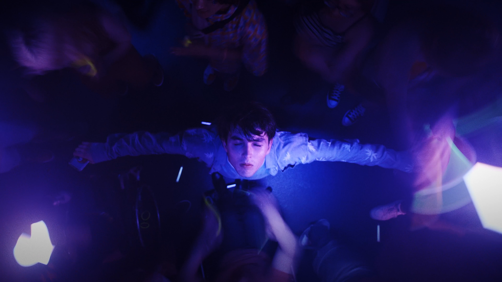 A medium shot from the film “DIVE DEEP” of a man facing upward with his eyes closed and his arms extended. The overhead lights produce a blue and purple hue in the room.
