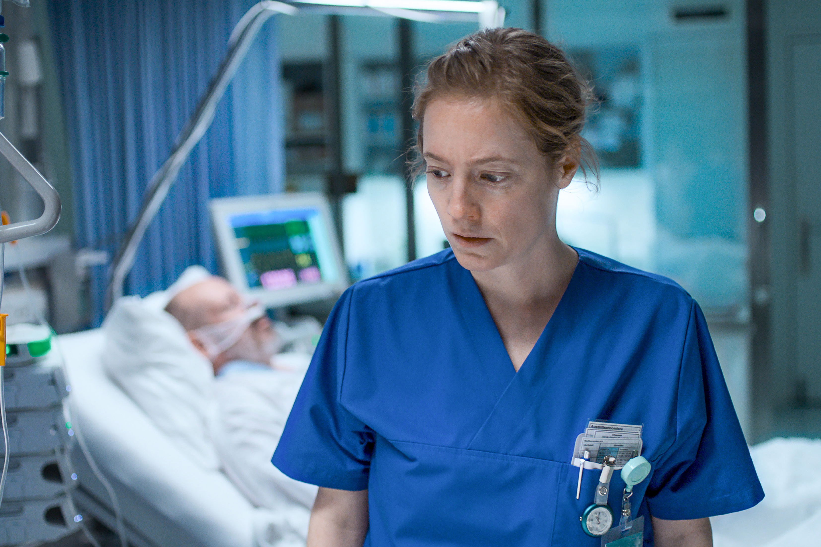 A medium shot from the film “Late Shift” of a woman wearing scrubs standing in front of a patient lying in a hospital bed.