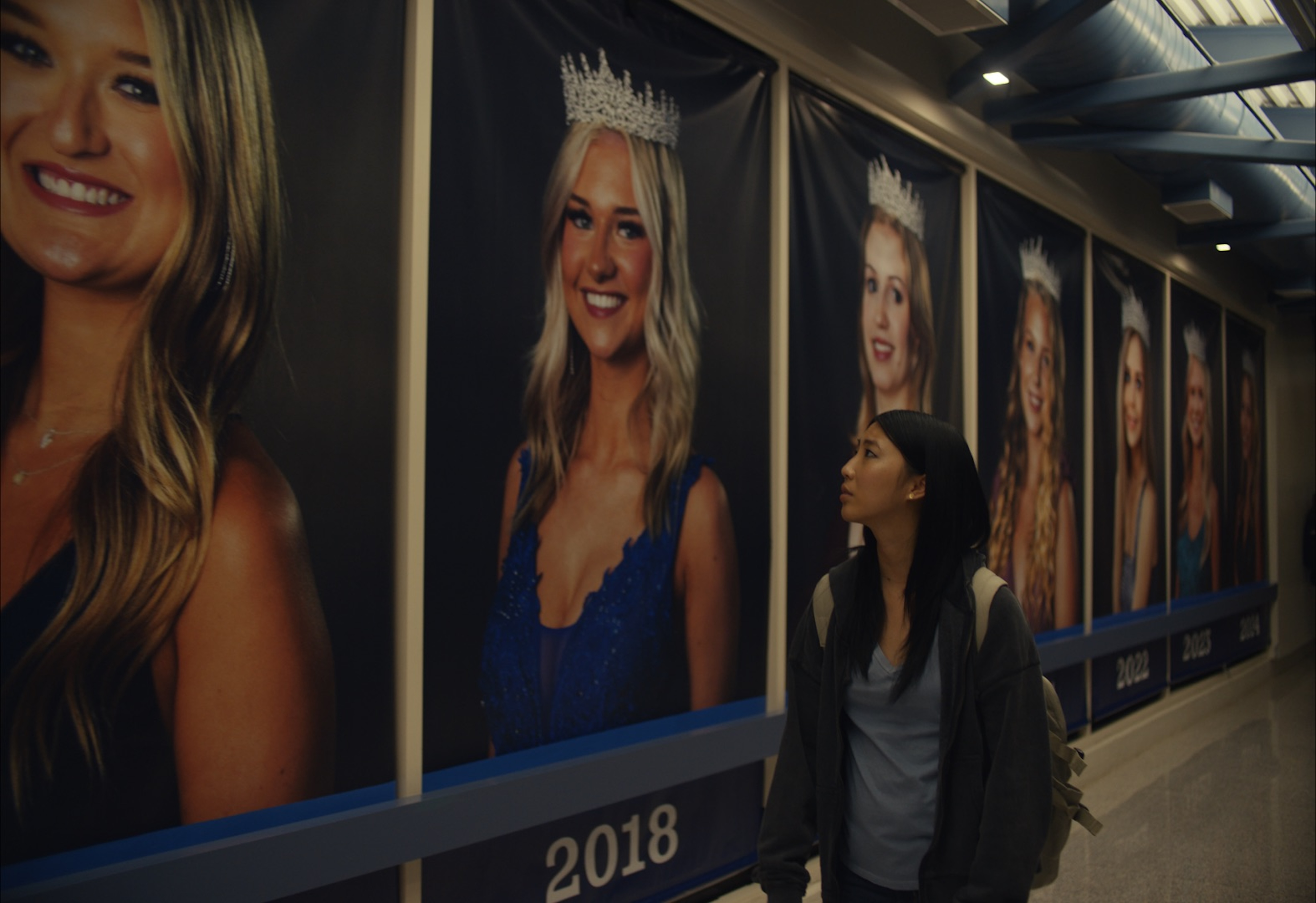 A wide shot from the film “Slanted” of a girl walking down a hallway lined with portraits of a high school’s previous prom queens.