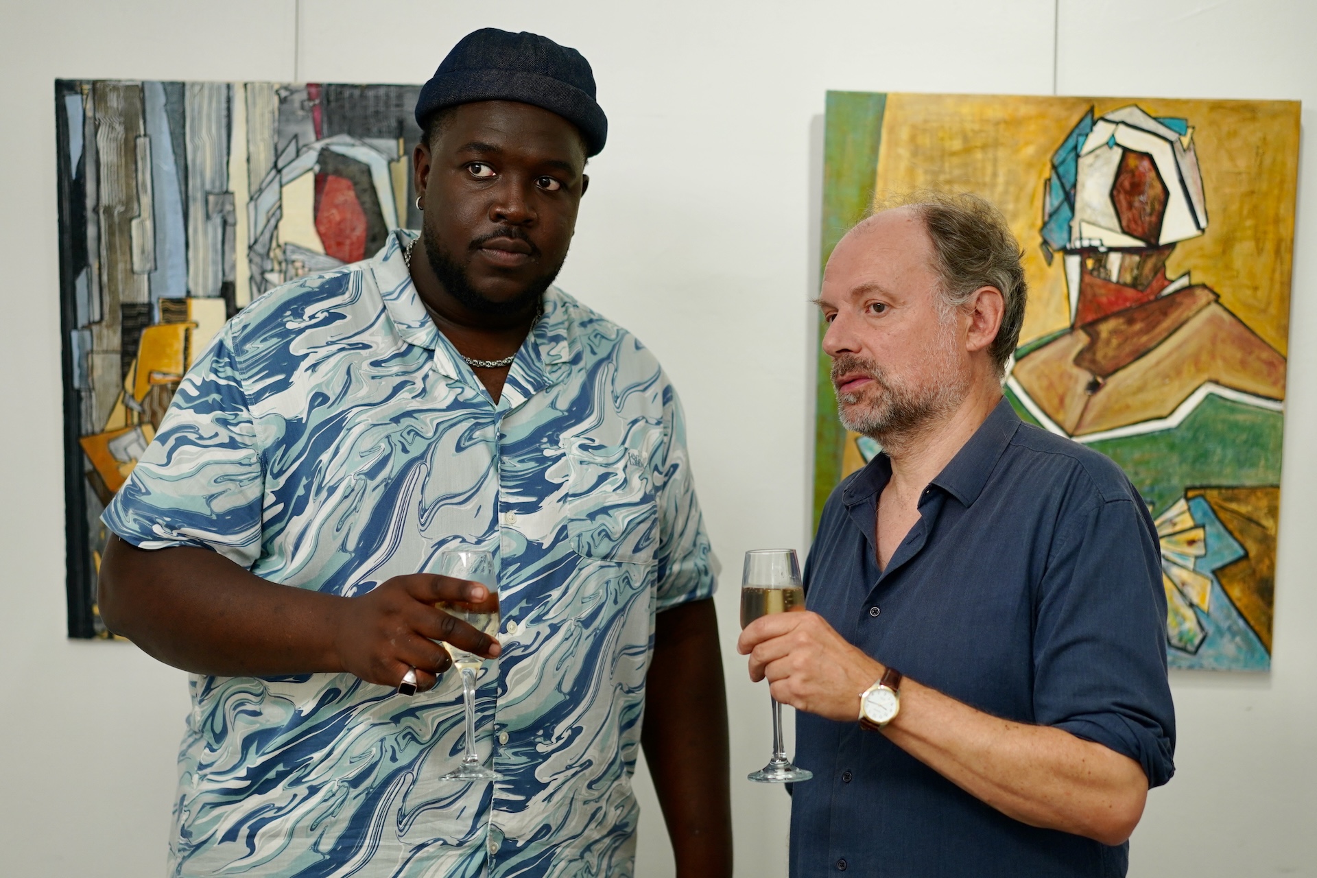 A medium shot from the film “Guess Who is Calling” of two men holding champagne glasses and standing in front of two art pieces.