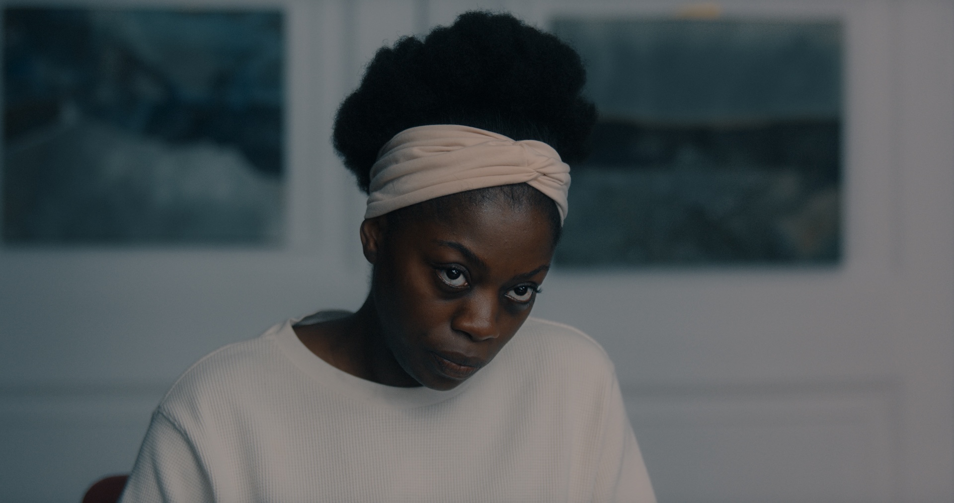 A medium shot from the film “MANGO” of a woman wearing a beige headband looking at the distance with a stern expression.