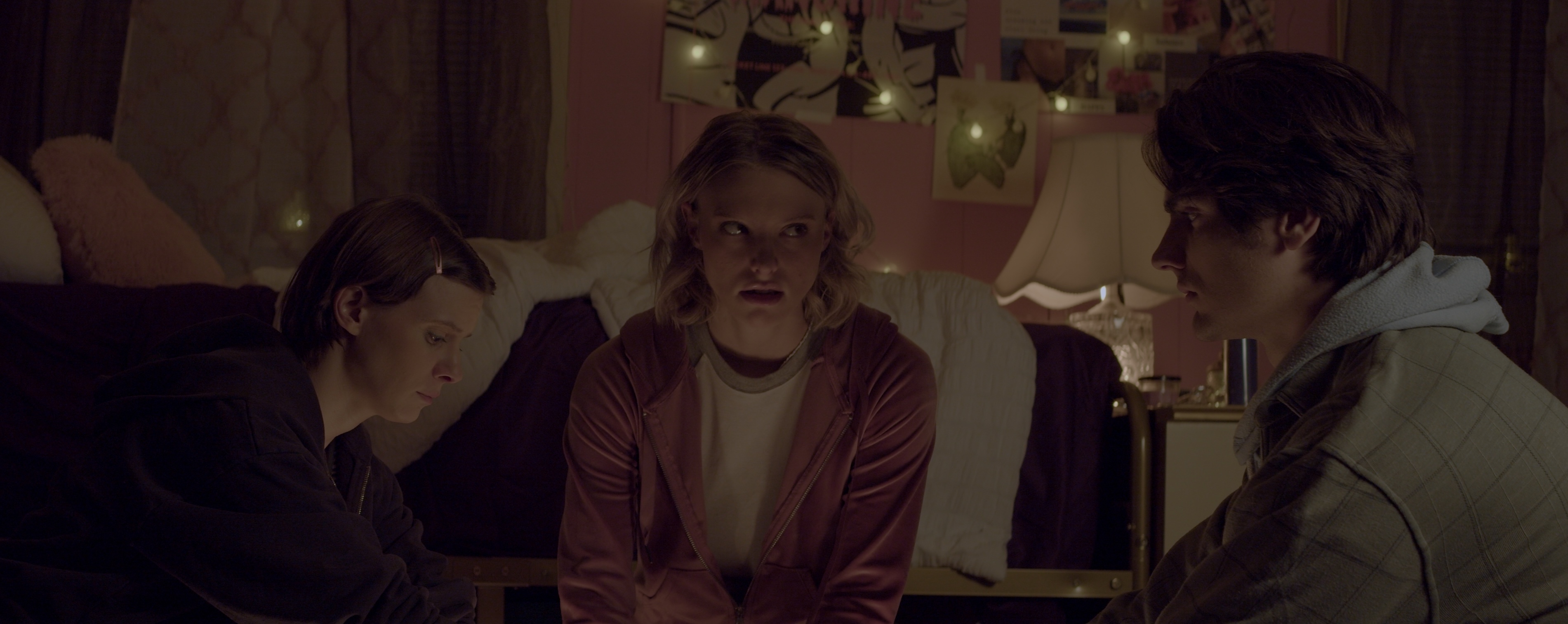 A medium shot from the film “Thrift Store Ouija Board” of three friends sitting in a dimly lit bedroom. One of the friends looks at the other with a serious glance.