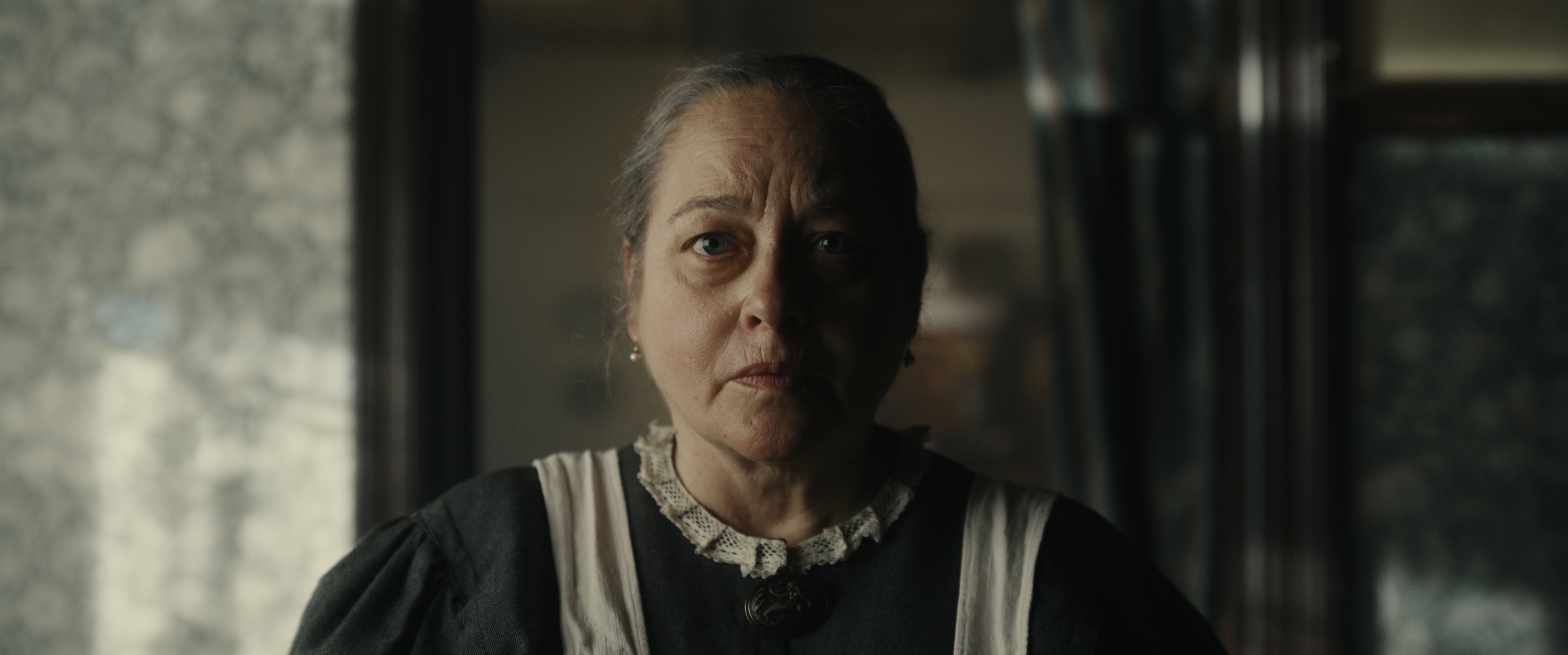 A close up shot from the film “The Pearl Comb” of a woman with slicked back hair looking forward in a dimly lit room.