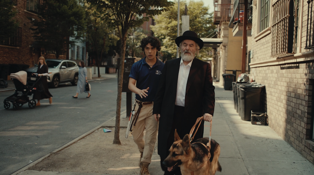 A medium shot from the film “Babka” of a blind Jewish Orthodox man walking his service dog and his guide trainer walking down a sidewalk.