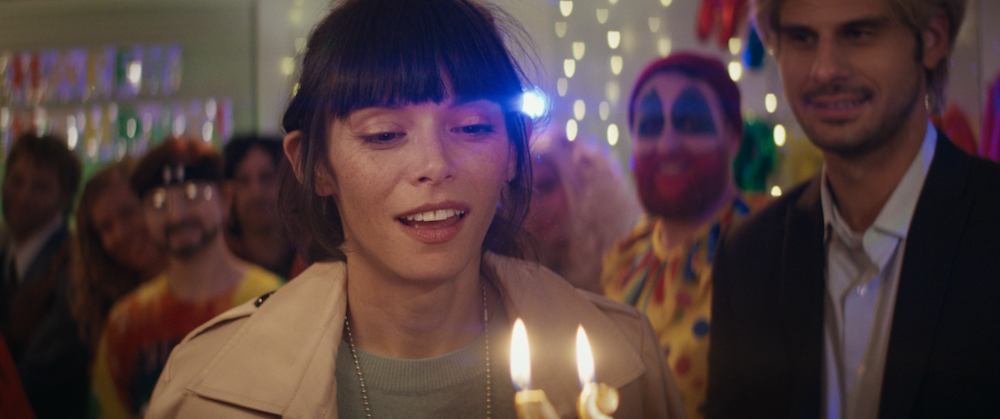 A medium shot from the film “My Favorite Murderer” of a woman looking at birthday candles. In the background there is a man dressed as a clown and “Happy Birthday” decorations.