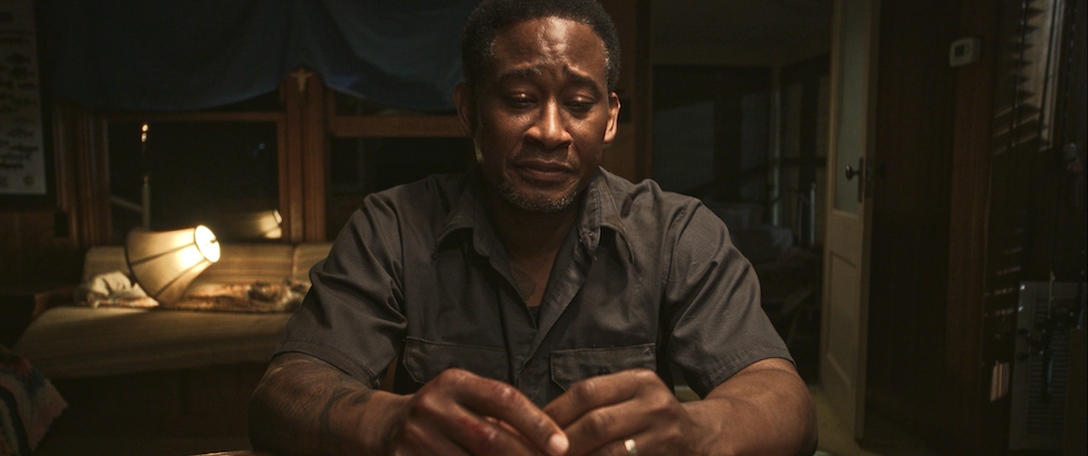 A medium shot from the film “Lake Placid Blue” of a man sitting with his fingers intertwined with a sad expression. The lamp in the background casts a warm light.