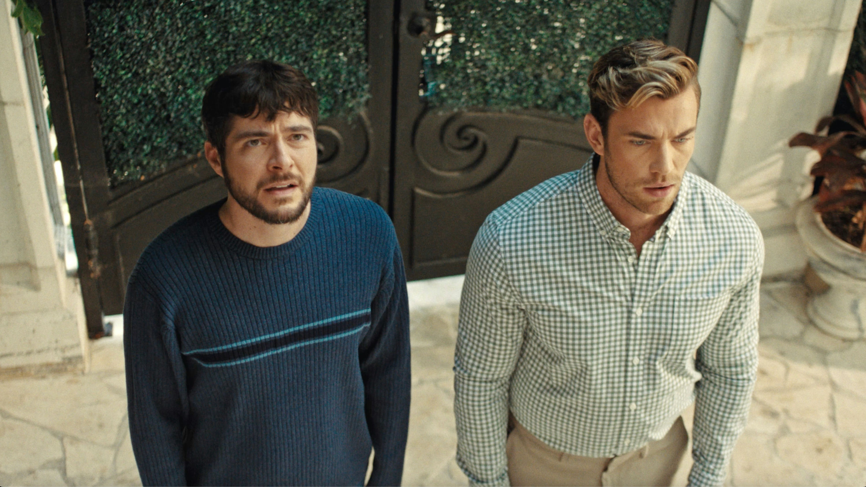 A medium shot from the film “She Raised Me” of two men standing in front of a gated entrance.