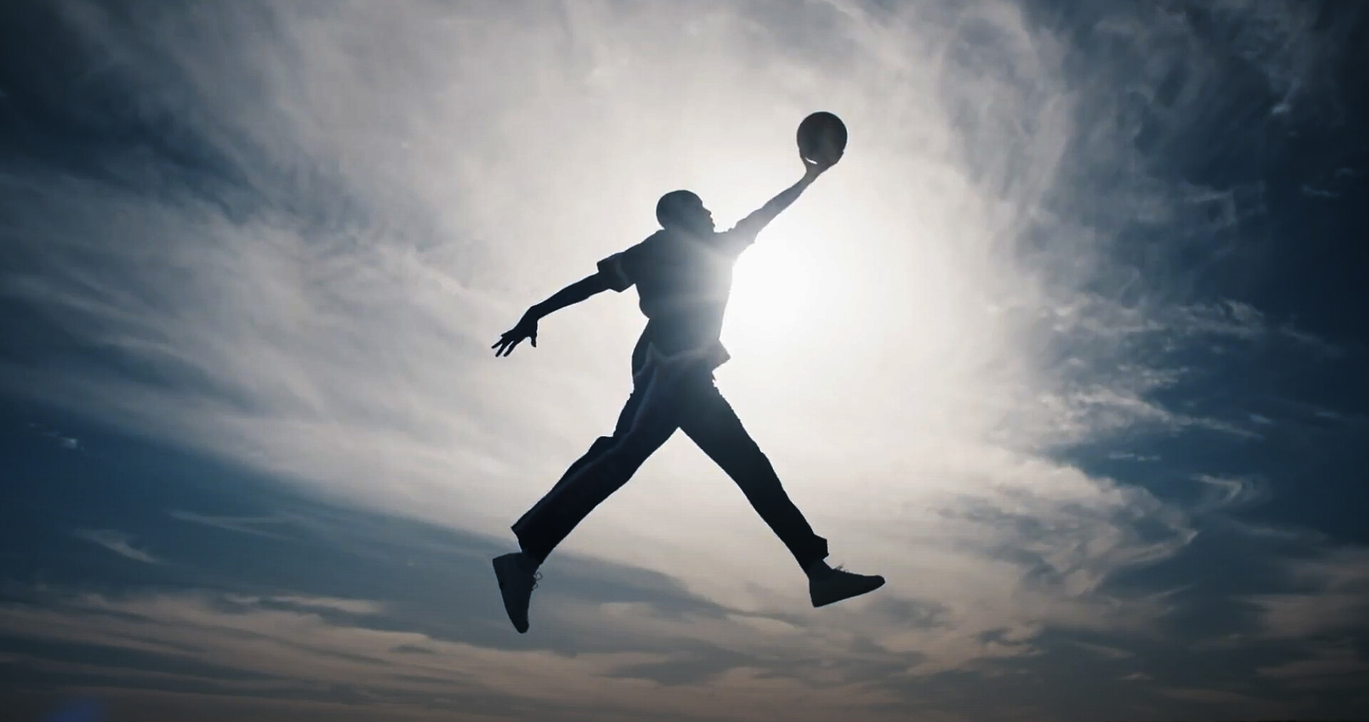 A wide shot from the film “Jumpman” of a man jumping into the air holding a ball, with the sun shining through him.