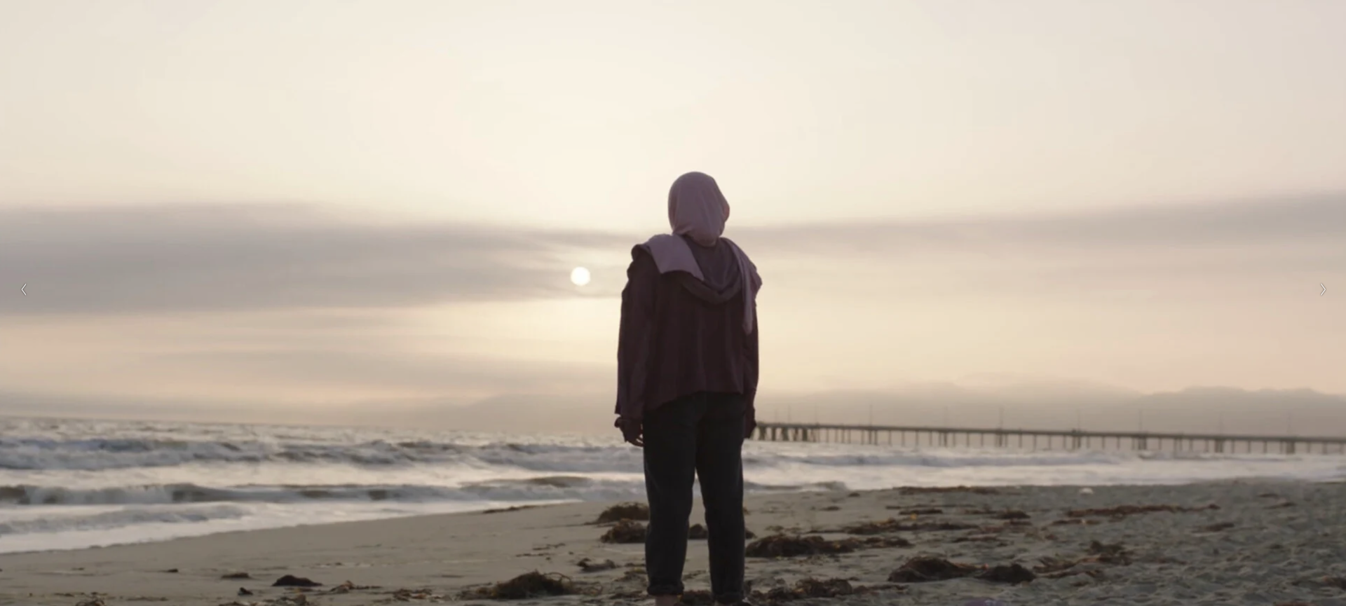 A wide shot from the film “Headbang” of a woman standing in the sand of a beach shore staring at the distant sun. A pier dock can be seen in the background.