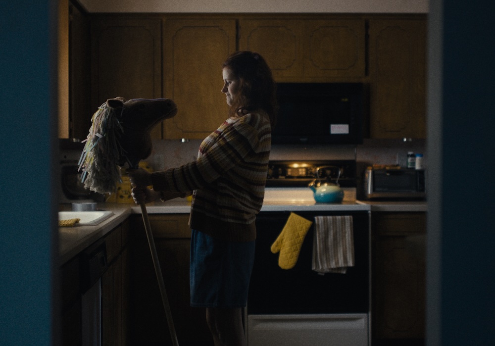 A medium shot from the film “Horsegirls” of a woman holding and looking at a hobby horse in the middle of her kitchen. The room is dimly lit with the exception of a warm light shining on the stovetop.