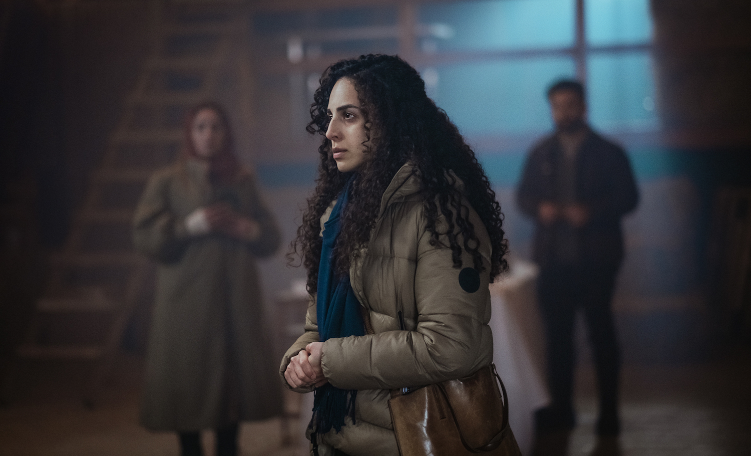 A medium shot from the film “Hatch” of a woman in a puffer jacket and a leather bag standing in a moonlit room.