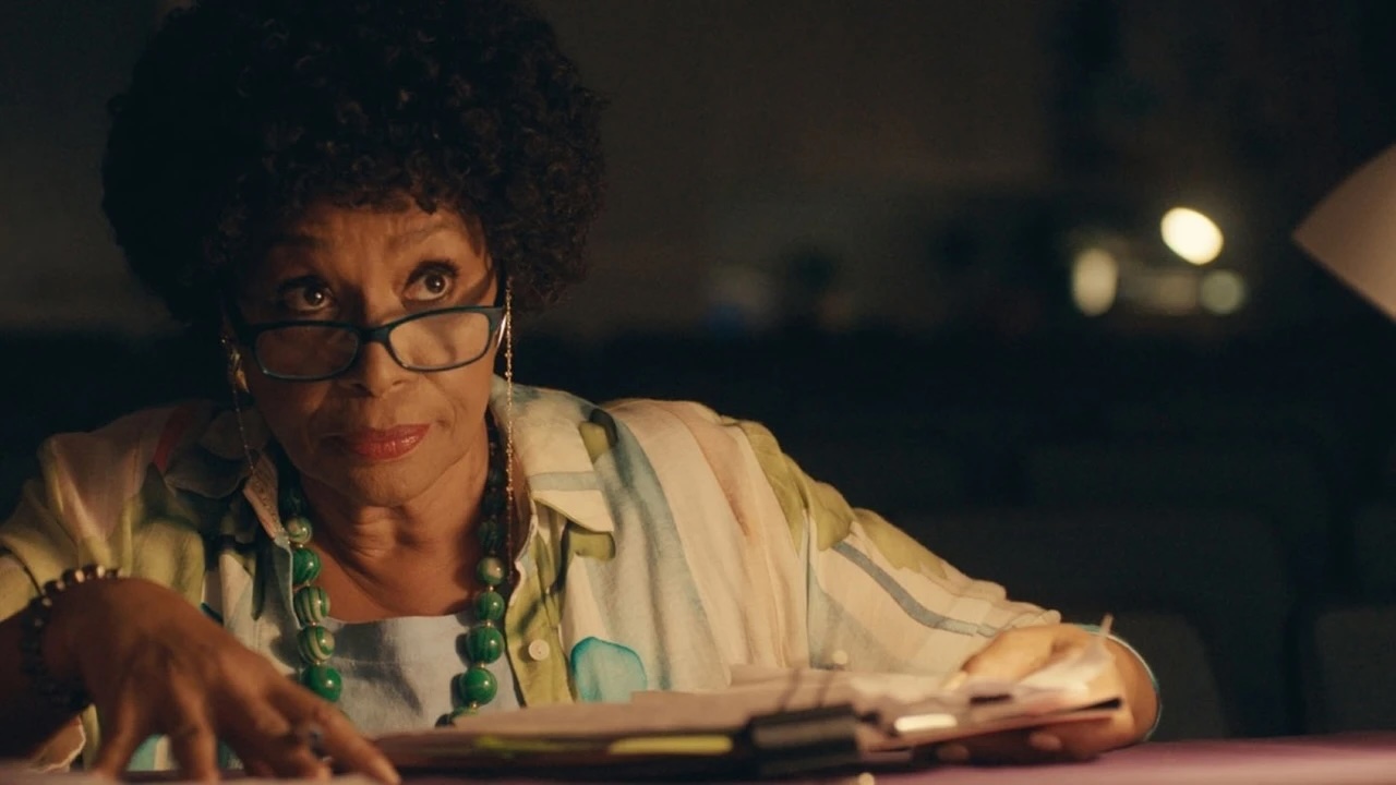 A medium shot from the film “Ado” of a woman in glasses studies and a clip board in warm lighting.