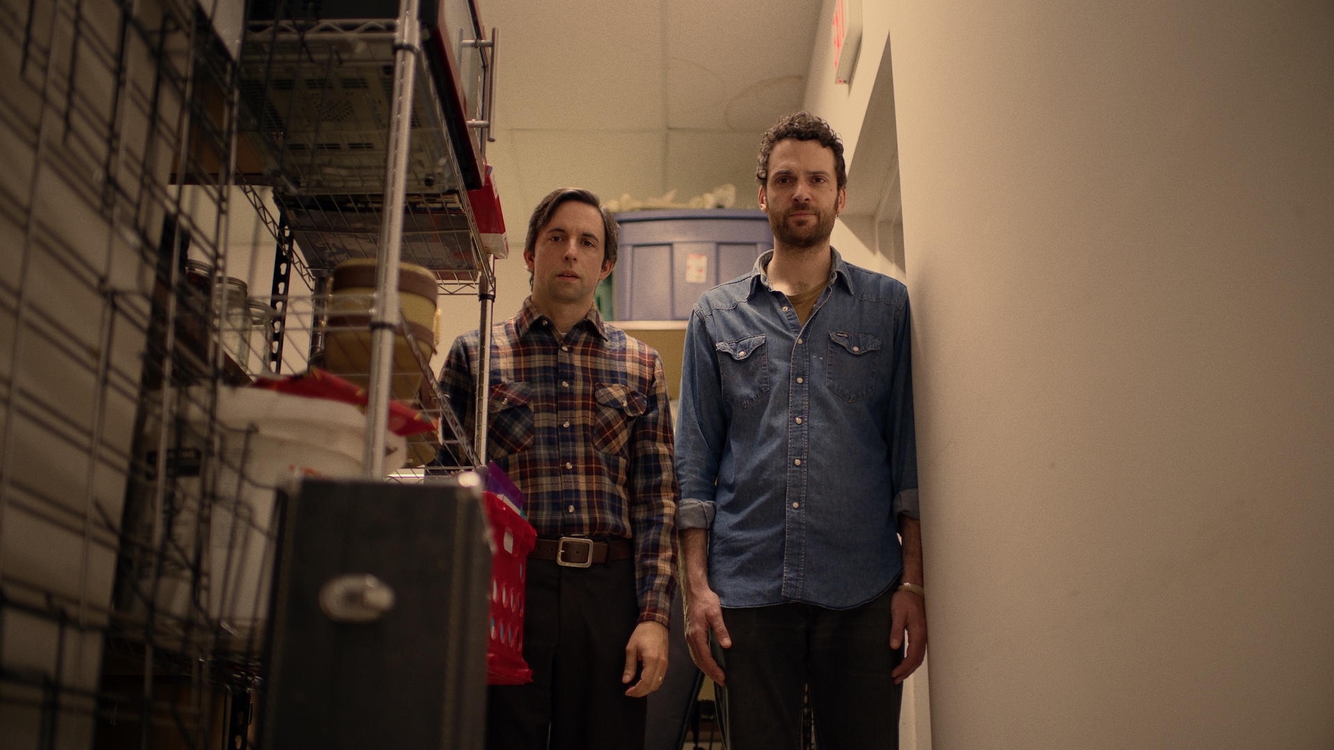 A medium shot from the film “The Music Store” of two men standing next to a metallic cabinet and looking at a musical instrument case.