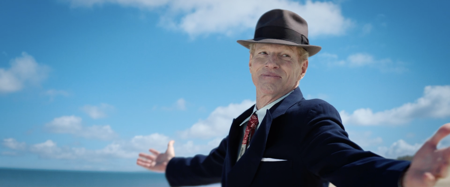 A medium shot from the film “The Ice Cream Man” of a man in a dark suit with his arms wide open. The sky is blue and clear.