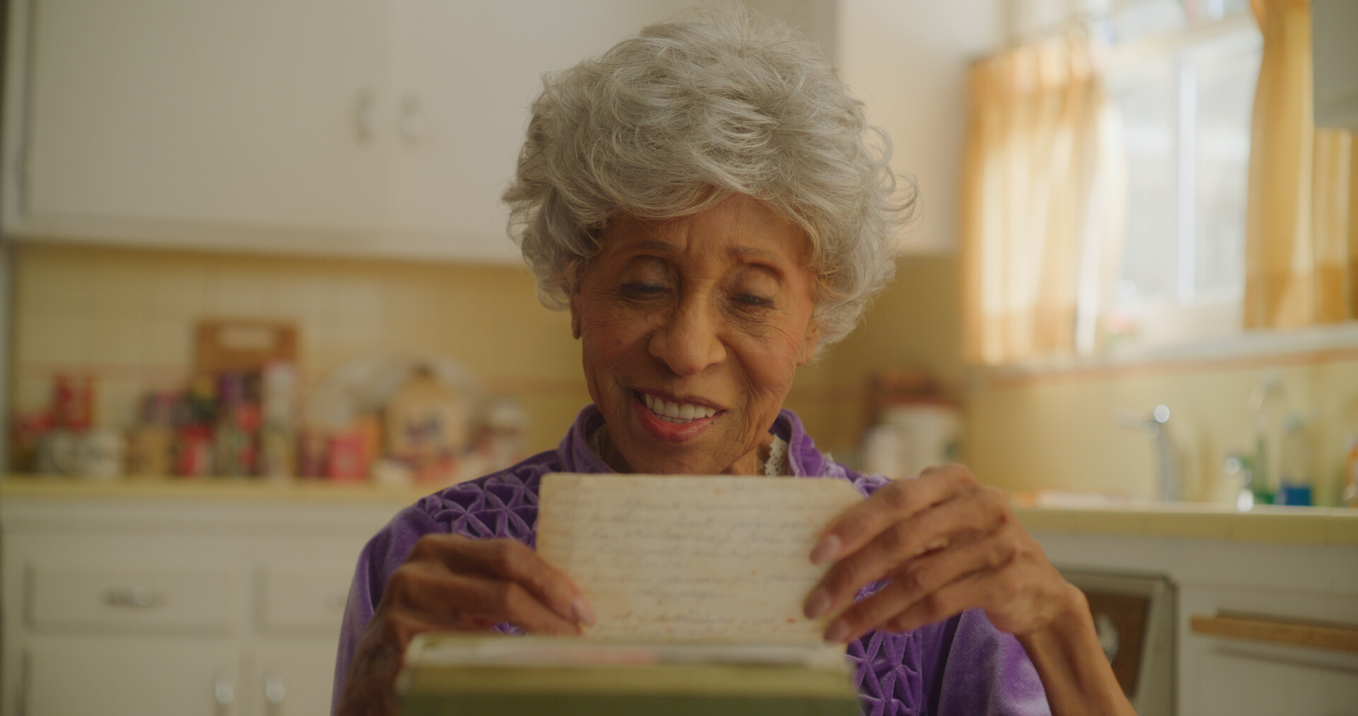 A close up shot from the film “Mildred 4 a Million” of a woman reading a note while sitting in her warmly lit kitchen.