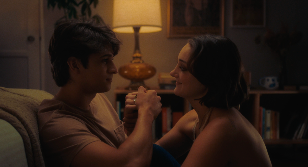 A medium shot from the film “All Roads Lead to Here” of a young couple holding hands. A lamp casts warm light from behind.