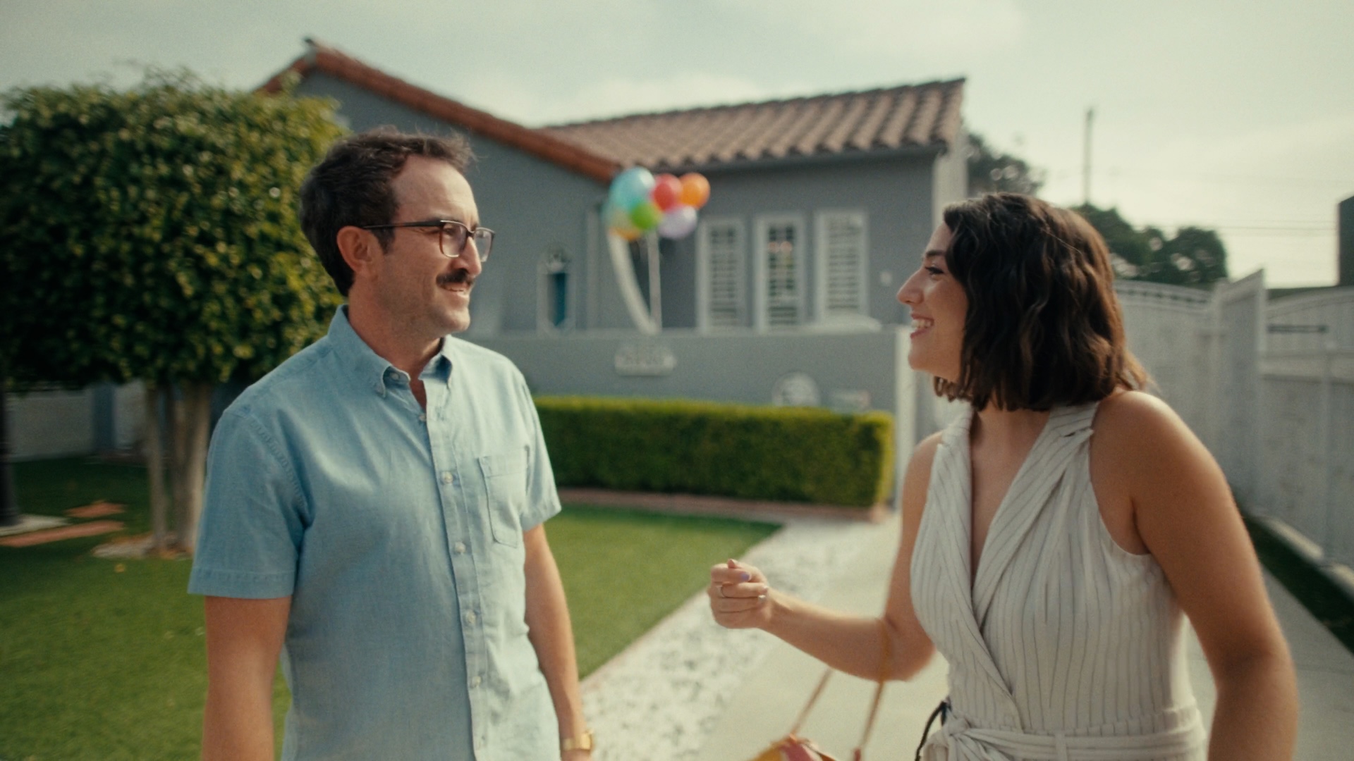 A medium shot from the film “Chips or Crackers” of a woman smiling at a man wearing glasses. A group of balloons floats in the distance.