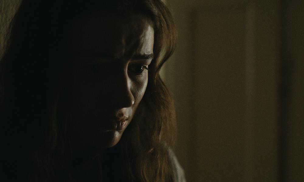 A close up shot from the film “Calf” of a woman staring at the distance scared in a dimly lit room, you can only see half of her face.