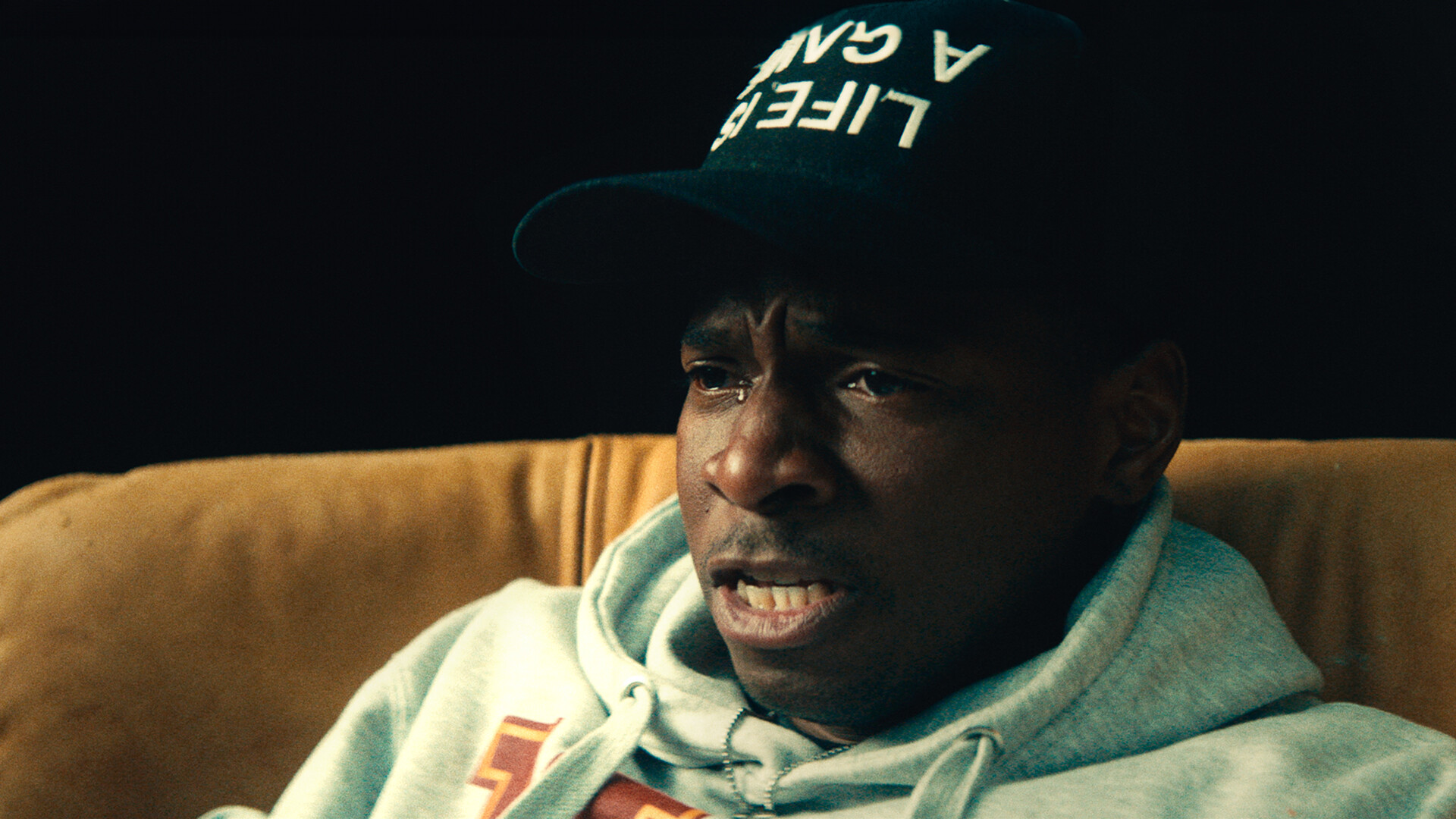 A close up shot from the film “Uncaged” of a man wearing a hoodie and a dark hat with a single tear running down his face.