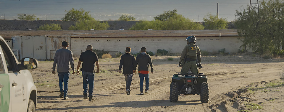A wide shot from the film “Beneath the Same Sky” of a group of four men being guided by a police officer on an all-terrain vehicle. The road is dusty and filled with tire tracks.
