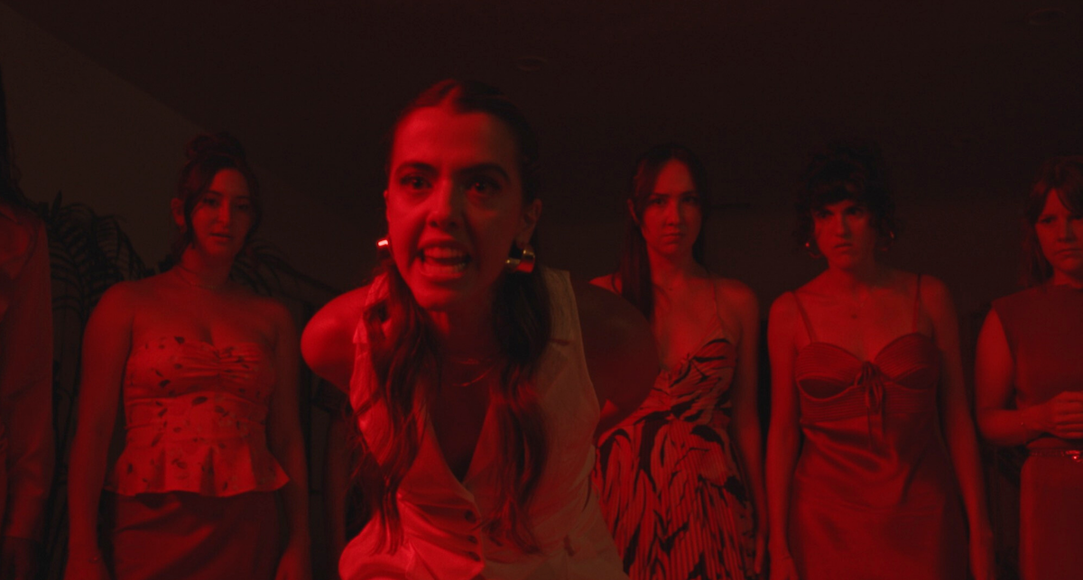 A medium shot from the film “She Devil” of a woman holding her hands behind her back with a mean expression. The other women stare intently at the object.