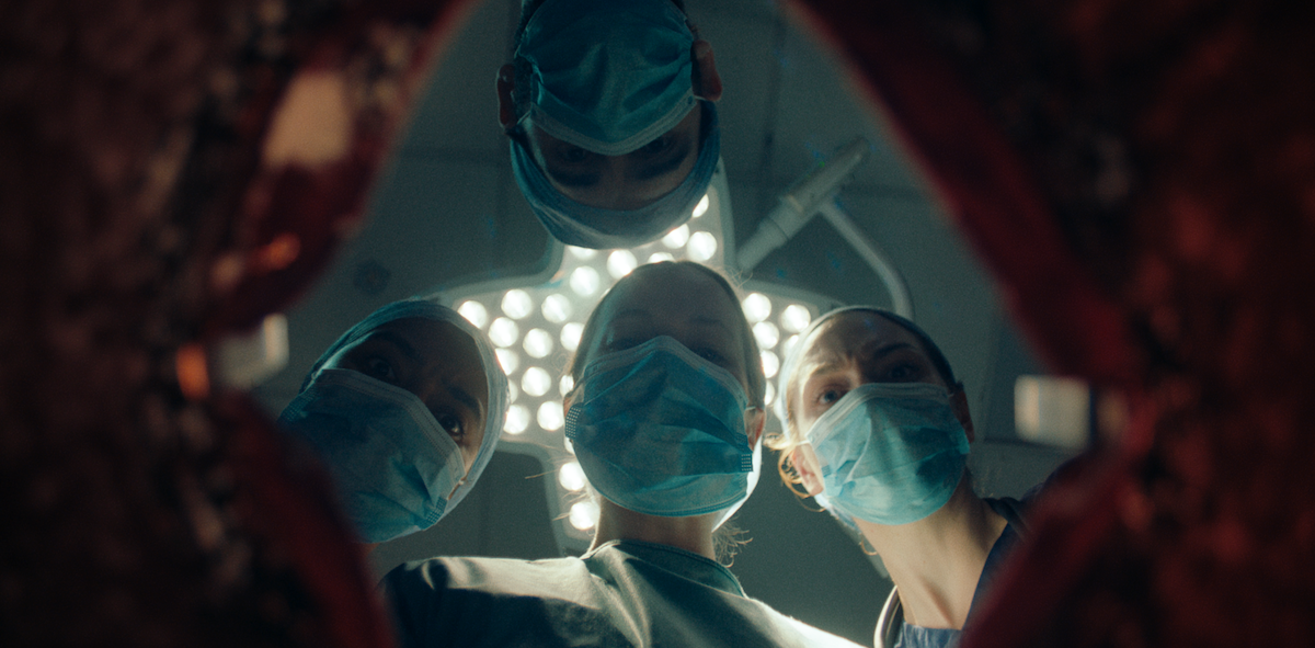 A medium shot from the film “Plastic Surgery” of four doctors wearing masks staring into a body opening of a patient. The overhead light casts a shadow on the doctors’ face.